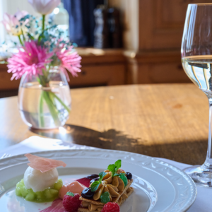 Ein Dessert mit frischen Beeren, Sahne und einer Kugel Sorbet steht auf einem Holztisch im JOURNALISTENCLUB, daneben ein Glas Weißwein und eine kleine Vase mit rosa und lila Blumen, die im Sonnenlicht liegen und die Skyline im Blick haben.