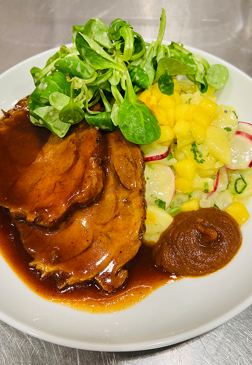 Gebratene Fleischscheiben mit brauner Bratensoße, serviert mit grünem Salat und Kartoffelsalat mit gelben Mangowürfeln, Radieschenscheiben und Kräutern auf einem weißen Teller - perfekt, um Ihre Kitchen Insights oder Ihr kulinarisches Feedback weiterzugeben.