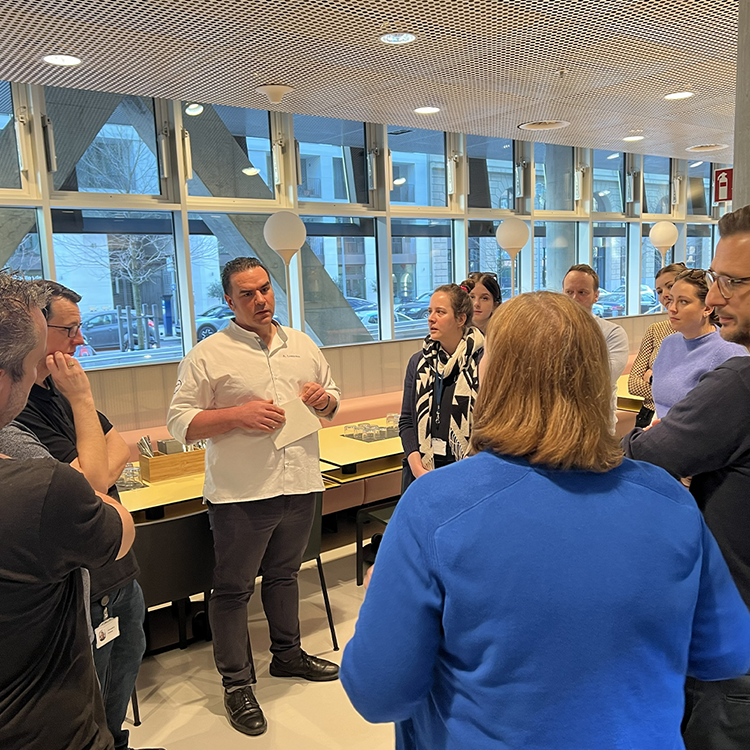 Eine Gruppe von Menschen steht in einem modernen Pausenraum eines Büros und hört aufmerksam einem Mann im weißen Hemd zu, der über Kitchen Insights berichtet. Im Hintergrund sind Tische, Stühle und große Fenster zu sehen, die eine Szene im Freien zeigen.
