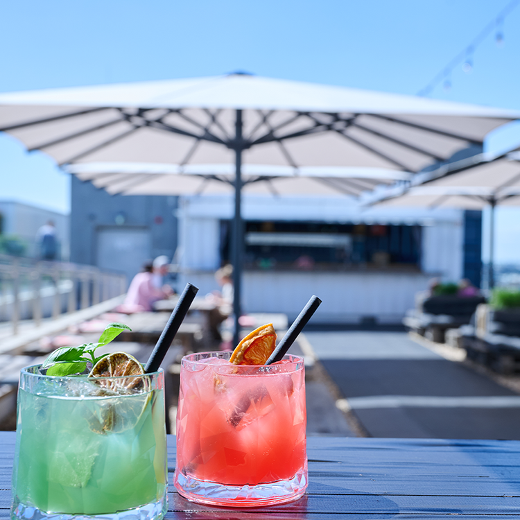 Zwei farbenfrohe Cocktails sitzen an einem sonnigen Tag auf einem Tisch unter großen weißen Terrassenschirmen an einer Bar im Freien - perfekt, um zu entspannen und neue Rezepte für den VEGANUARY 2026 zu finden. Der eine Drink ist grün mit Limette und Minze, der andere pink mit getrockneter Orange.