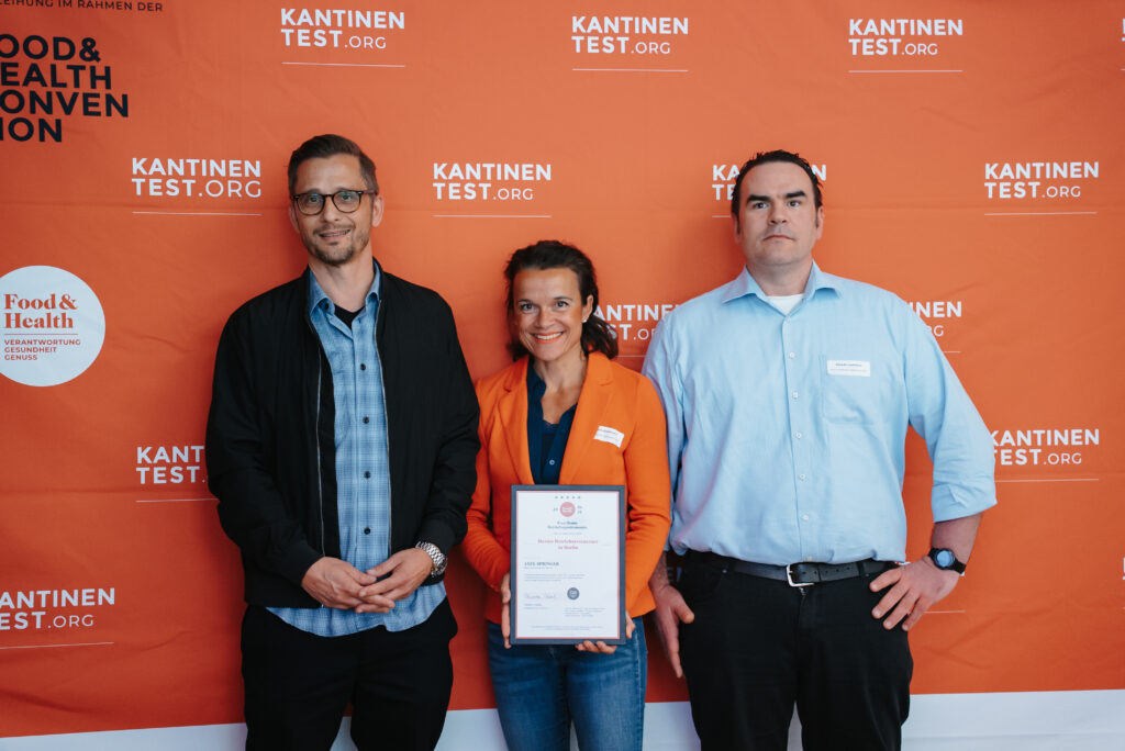 Three people are standing in front of an orange background featuring the logos of Food & Health Kantinentest and the company. The person in the middle is holding a framed certificate and smiling, while the others stand on either side, looking at the camera.