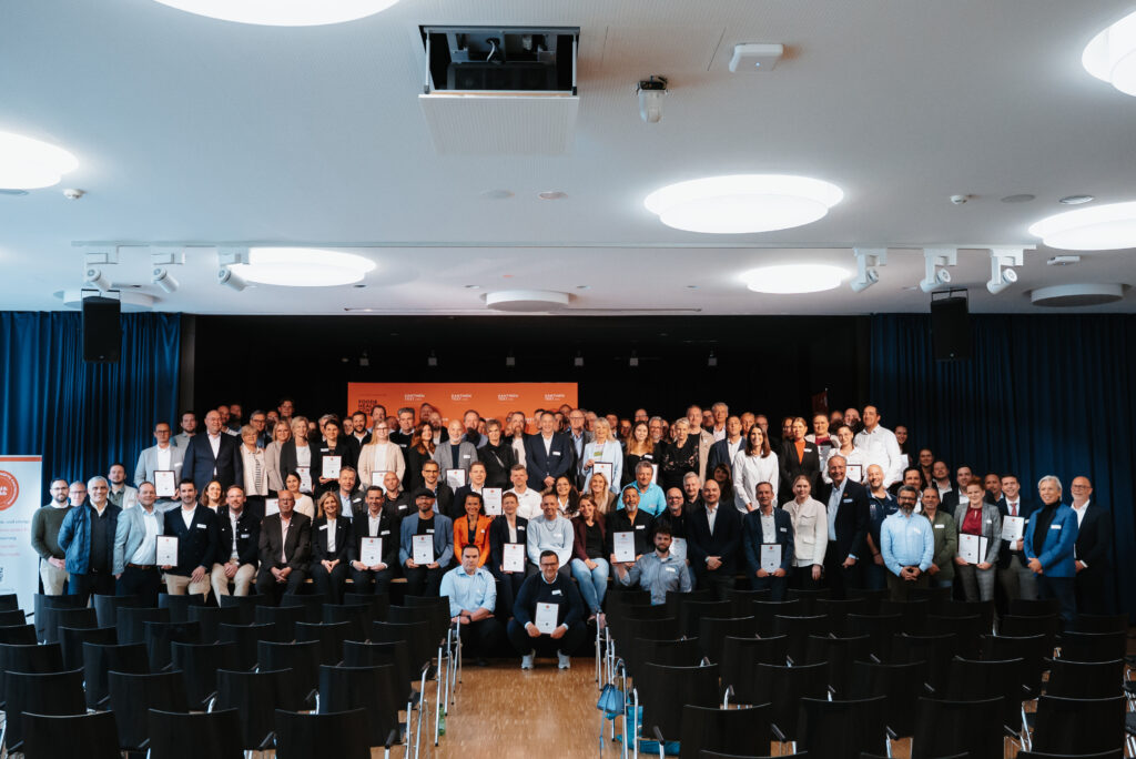 A large group of people is posing together in front of an auditorium, some holding certificates for the Food & Health cafeteria test, with empty rows of seats in front of them and a stage with a red backdrop behind them.