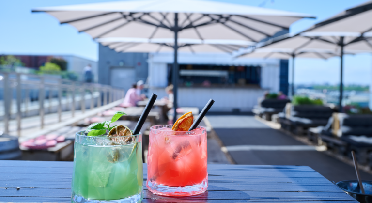 Zwei farbenfrohe Cocktails, ein grüner und ein rosafarbener, stehen auf einem Tisch unter großen weißen Sonnenschirmen in einer sonnigen Dachterrassenbar während der REM-Veranstaltungen, mit unscharfen Sitzplätzen und Menschen im Hintergrund.
