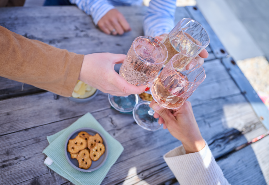 Drei Personen stoßen mit Weingläsern über einem rustikalen Holztisch mit Snacks wie Brezeln und Käse im Freien an. Die Szene zeigt ein zwangloses REM Events-Treffen, perfekt, um gemeinsam den Saisonstart zu feiern.