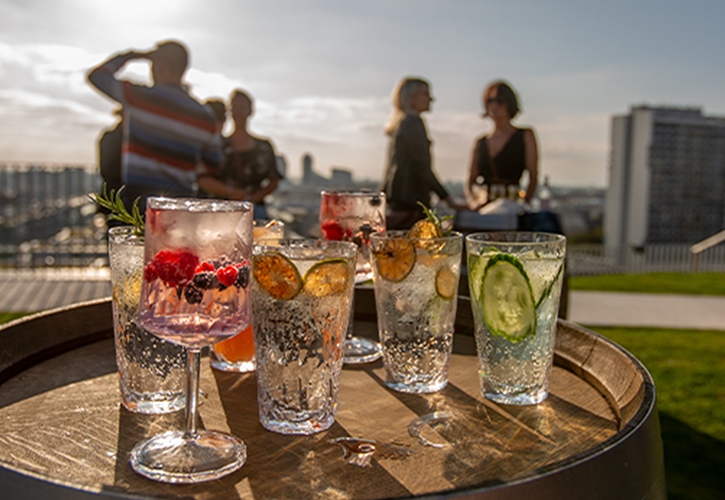Bei einem sonnendurchfluteten REM-Event im Freien werden sechs bunte Cocktails auf einem Holzfass serviert, garniert mit Früchten und Kräutern, während vier Personen im unscharfen Hintergrund vor den Gebäuden der Stadt Kontakte knüpfen und einen lebhaften Saisonstart markieren.