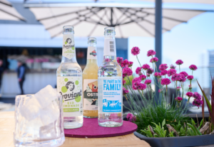 Drei Flaschen Limonade stehen auf einem Holztisch neben einem Glas Eis und rosafarbenen Blumen, mit großen weißen Terrassenschirmen und einem modernen Gebäude im Hintergrund - perfekt, um den Saisonstart bei REM Events zu feiern.