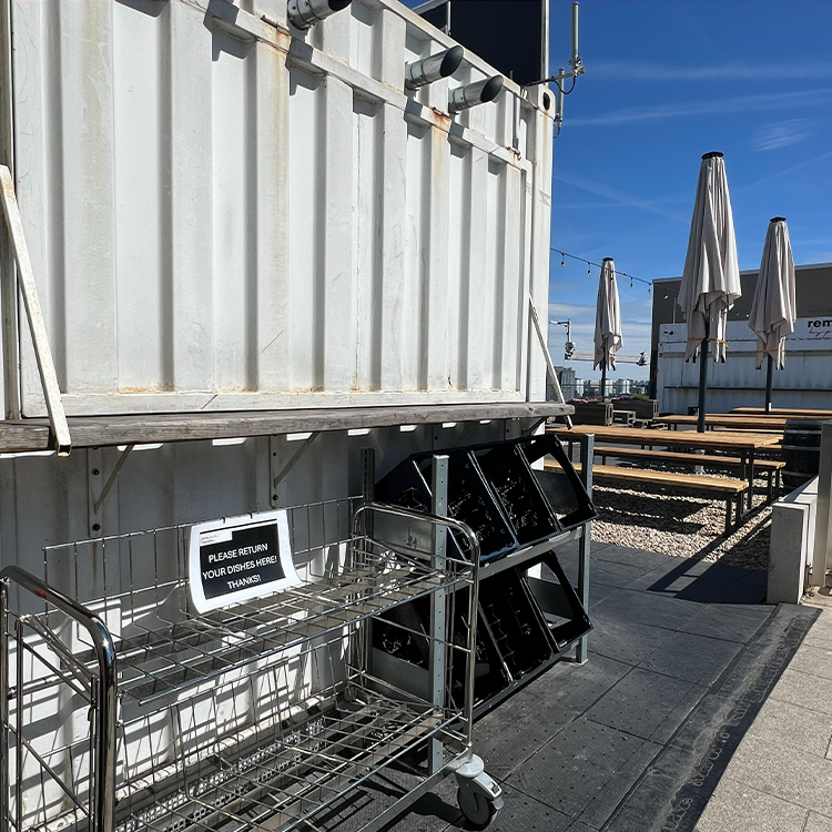 Sitzbereich im Freien mit geschlossenen Sonnenschirmen, Holzbänken und Kiesboden. Ein Metallwagen mit der Aufschrift "Bitte geben Sie Ihre TASSEN zurück, wenn Sie fertig sind" steht neben einem weißen Container unter einem klaren blauen Himmel.