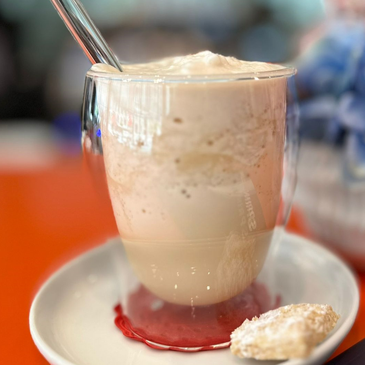 Eine Glastasse mit schaumigem Eiskaffee und einem Glasstrohhalm steht auf einer weißen Untertasse, begleitet von einem gepuderten Keks, auf einem orangefarbenen Tisch. Diese erfrischende Sommergetränkeszene steht vor einem schön verschwommenen Hintergrund.
