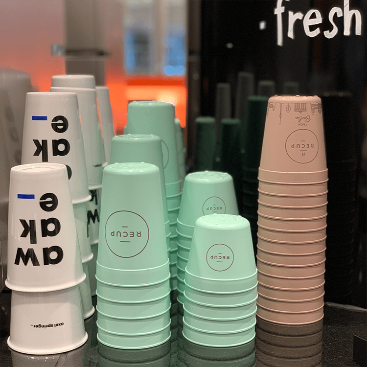 Stapel von nachhaltigen, wiederverwendbaren Bechern in Pastellfarben, darunter Weiß, Mintgrün und Hellrosa, sind ordentlich auf einer Arbeitsplatte vor einem schwarz-orangen Hintergrund angeordnet - perfekt für deinen Kaffee oder Lunch. Auf einigen Bechern steht das Wort RECUP.