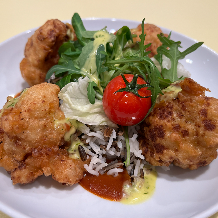 Ein Teller mit knusprig gebratenen Hähnchenteilen, Wildreis, einer Kirschtomate, gemischtem Grün und Salat, beträufelt mit cremigen und roten Saucen - perfekt, um ihn zu genießen, bevor man sich auf die Suche nach Tassen mit einem lustigen Spruch im Schrank macht.