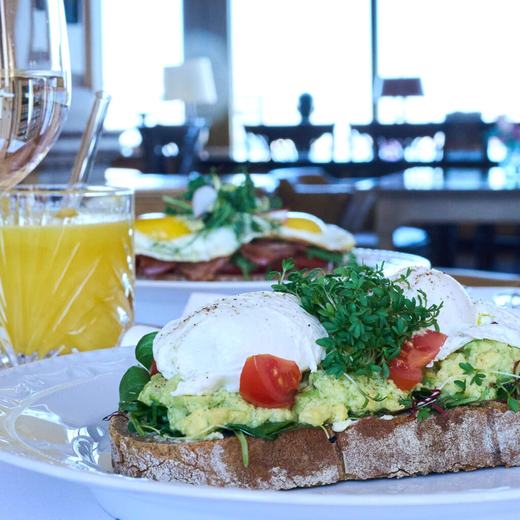 Ein Teller mit Toast, belegt mit Avocado, pochierten Eiern, in Scheiben geschnittenen Tomaten und frischem Grün, daneben ein Glas Orangensaft. Ein weiterer Teller mit Eiern und Fleisch steht unscharf im Hintergrund.