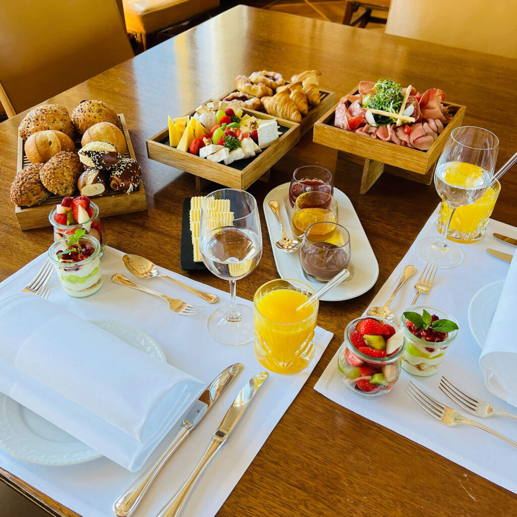 Ein gedeckter Frühstückstisch mit Gebäck, Brötchen, Käse, Fleisch, Obstparfaits mit Erdbeeren, Gläsern mit Orangensaft und Geschirr auf weißen Tischsets. Die Speisen sind ordentlich auf Holztabletts und -tellern angerichtet.