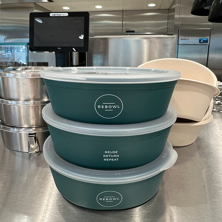 Drei grüne RE-BOWL Lunchbehälter mit dem Slogan "Reuse Return Repeat" stehen nachhaltig gestapelt auf einer Edelstahltheke in einer Profiküche, neben gestapelten Metall- und beige Containern.