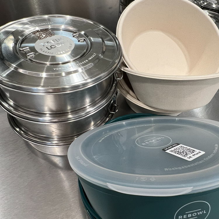 Ein Stapel Lunchboxen aus Metall, zwei beigefarbene kompostierbare Schüsseln und ein dunkelgrüner wiederverwendbarer Behälter mit durchsichtigem Deckel stehen auf einer Edelstahlfläche - fast bereit für den Schrank oder für Ihre Lieblingstassen.
