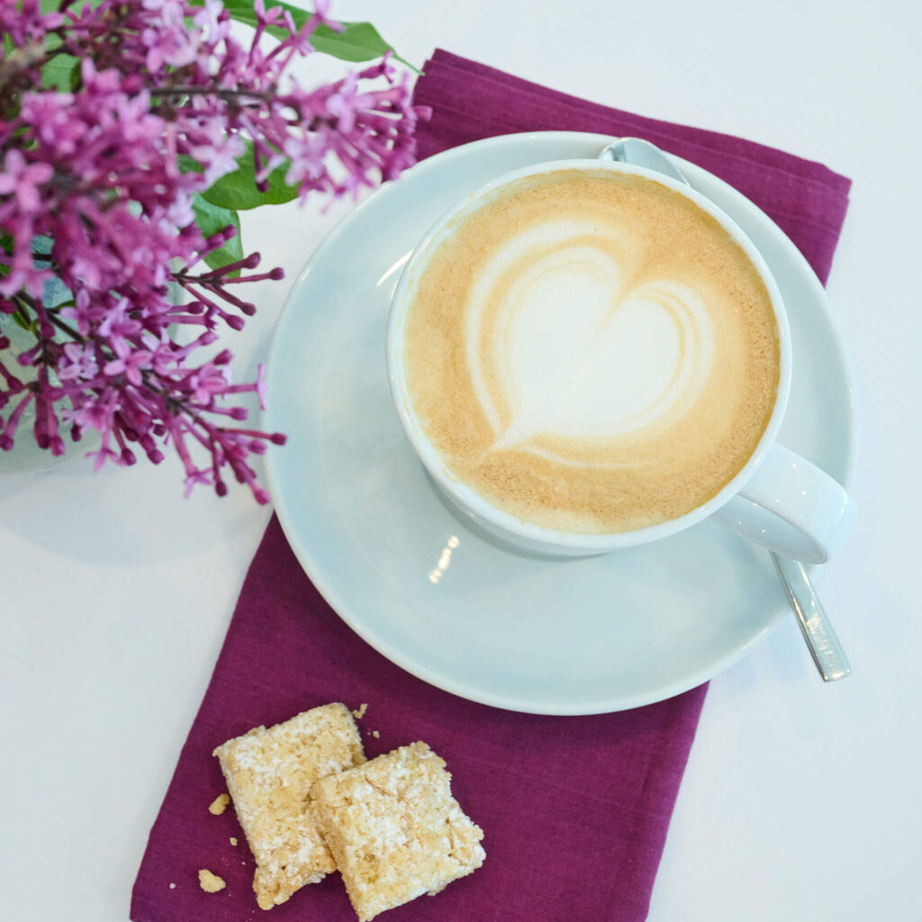 Eine Tasse Kaffee mit herzförmiger Latte-Art auf einer weißen Untertasse, die auf einer violetten Serviette neben zwei mürben Keksen und einem Strauß violetter Blumen steht.