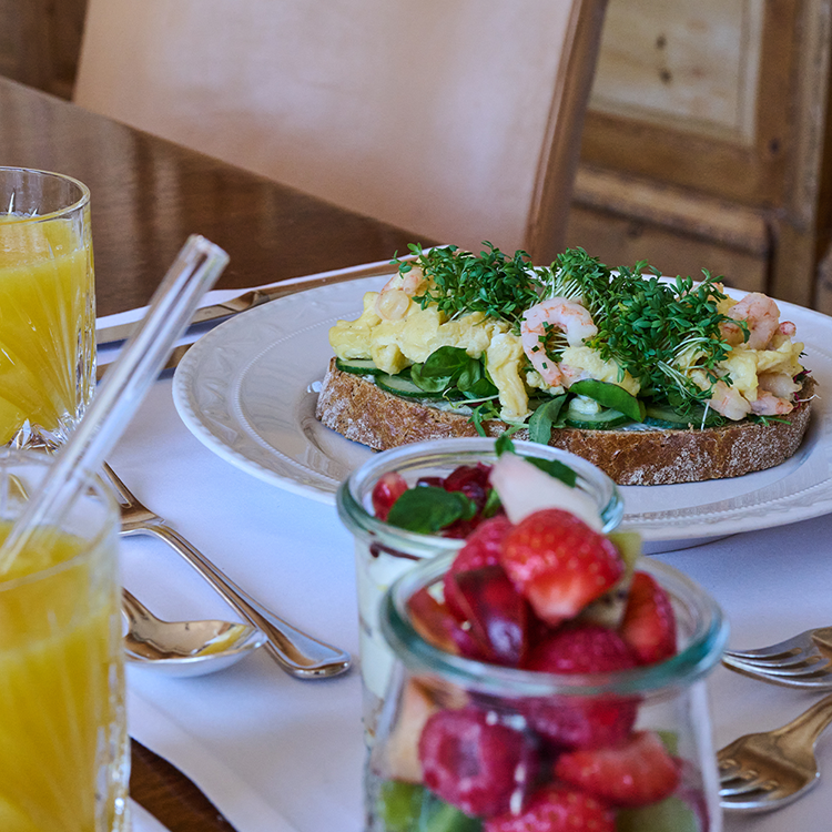 Ein zum Frühstück gedeckter Tisch mit Gläsern voller Orangensaft, einem Teller mit offenen Sandwiches, belegt mit Rührei, Blattgemüse und Garnelen – dazu Gläser mit frischem Obstsalat – lädt dazu ein, die Mahlzeit in gemächlichem Tempo zu beginnen.