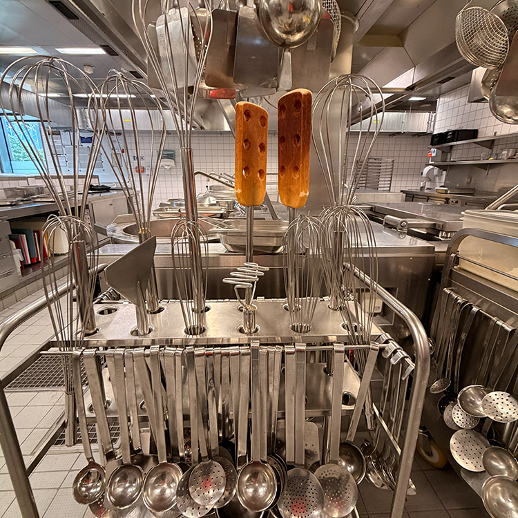 Eine Großküche mit Metallutensilien wie Schneebesen, Schöpfkellen und Löffeln. Zwei rechteckige Brotlaibe sind auf Schneebesen aufgespießt und stehen aufrecht in der Mitte zwischen den Utensilien - wahre Küchenerkenntnisse in Aktion.
