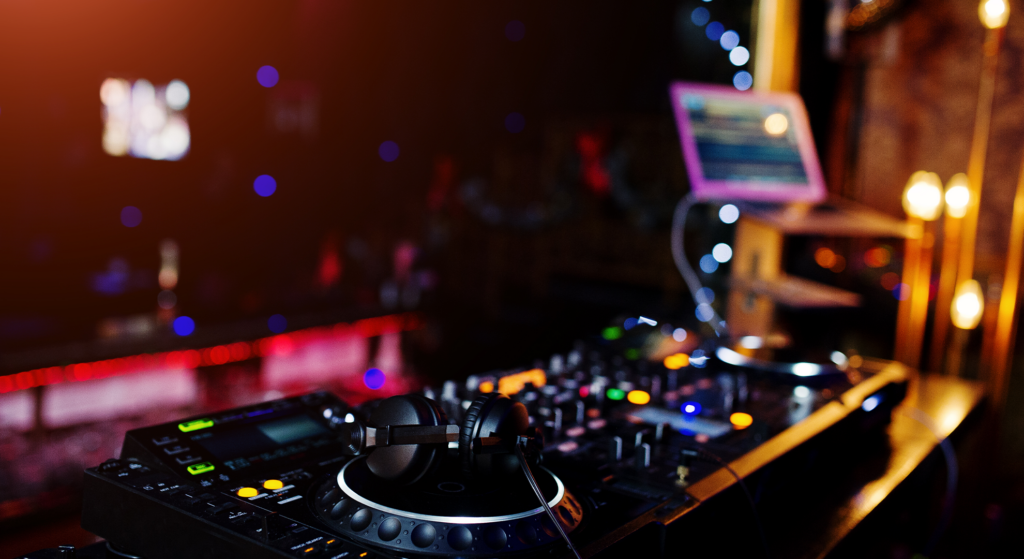 Eine Nahaufnahme eines DJ-Mischpults mit Kopfhörern, die auf einem Plattenteller liegen, bunte Lichter und ein Laptop-Bildschirm im Hintergrund bilden die Bühne - Bühne frei - für REM Talente, die in einer lebhaften Nachtclub-Atmosphäre glänzen.