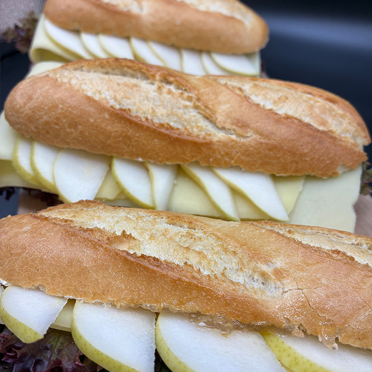 Drei Baguette-Sandwiches, gefüllt mit Käsescheiben und frischer Birne, nebeneinander aufgereiht. Das Brot hat eine knusprige, goldbraune Kruste, und die Zutaten sind in jedem Sandwich ordentlich geschichtet.