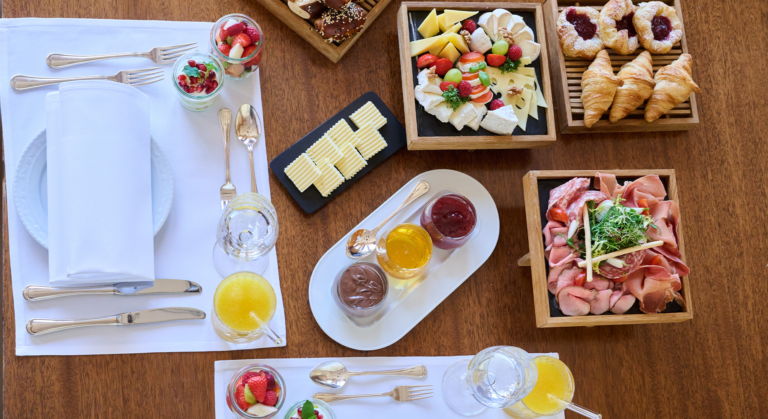 Ein reichhaltiges Frühstück auf einem Holztisch gibt den Ton für den Tag an – Frühstücken mit Gebäck, Croissants, Käse, Obst, Aufschnitt mit Salat, Butter, Konfitüre, Orangensaft und Joghurt-Parfaits – die schönste Mahlzeit, die man mit Besteck und Servietten genießt.