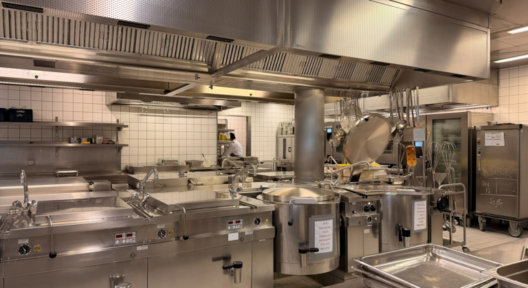 Eine große Großküche aus rostfreiem Stahl mit Industriegeräten, Töpfen und Pfannen. Der Raum ist sauber und organisiert, mit weiß gefliesten Wänden und einer Person, die im Hintergrund arbeitet - perfekt für KITCHEN INSIGHTS oder JOURNALISTENCLUB-Veranstaltungen.