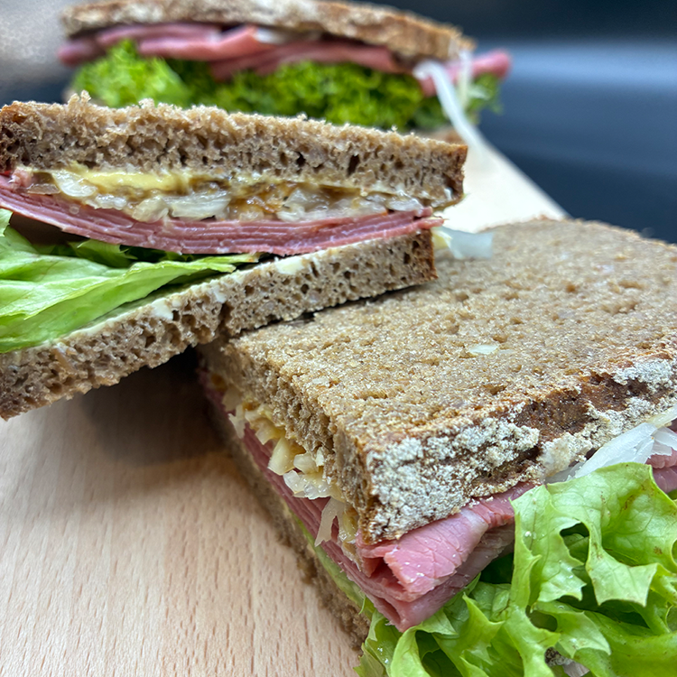 A close-up of two rye bread sandwiches stacked on top of each other with lettuce, cold cuts, onions, and melted cheese on a wooden board, with the sandwich in the foreground cut in half.