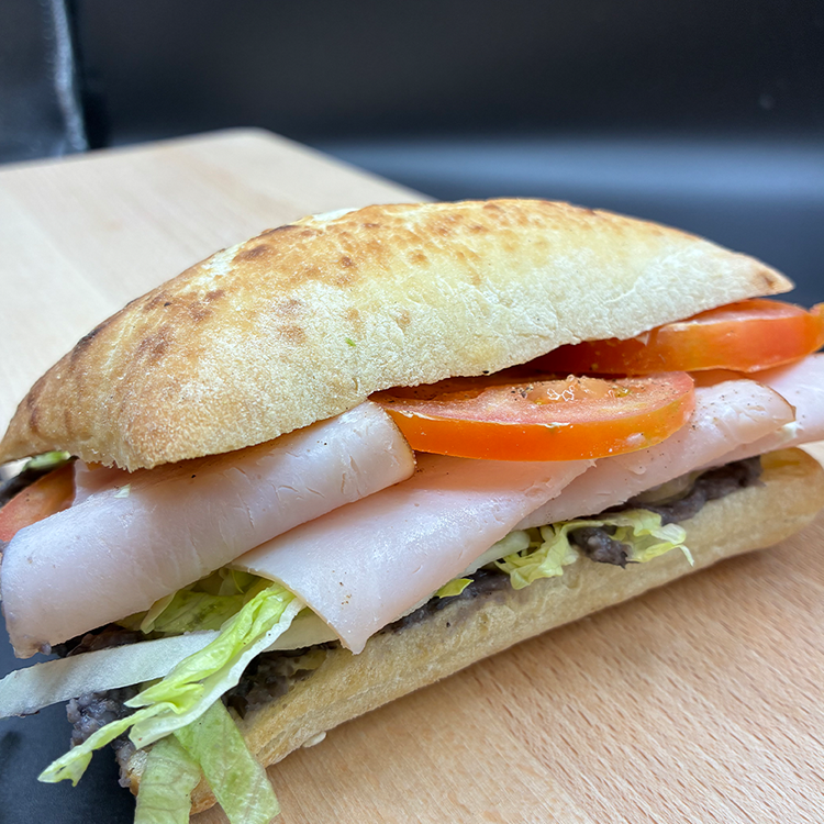 A sandwich on ciabatta bread, filled with slices of turkey breast, tomatoes, shredded lettuce, and a black bean spread, served on a wooden cutting board.