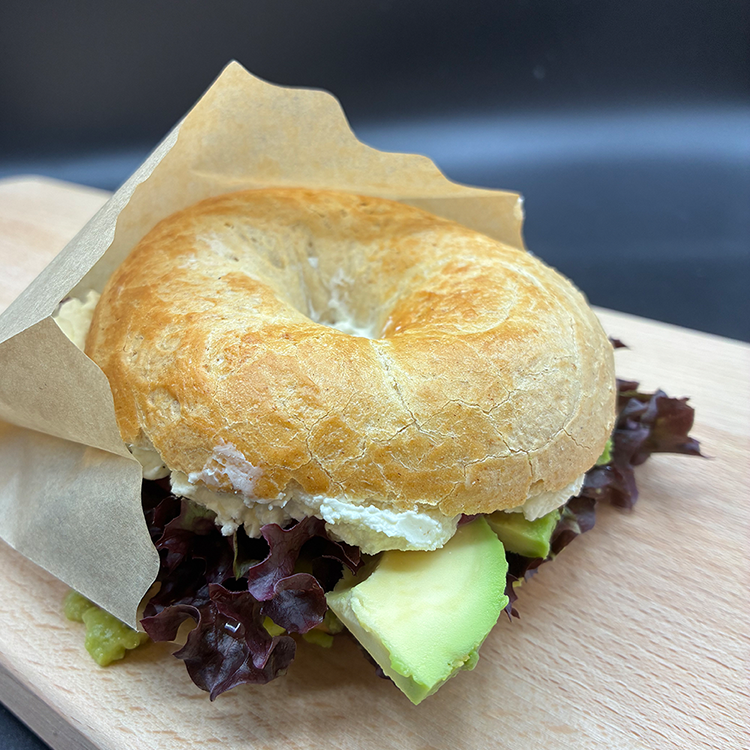 A bagel sandwich filled with lettuce, avocado slices, and cream cheese—partially wrapped in brown parchment paper—rests on a wooden board against a dark background, ready to take you on a culinary journey around the world at the DELI.