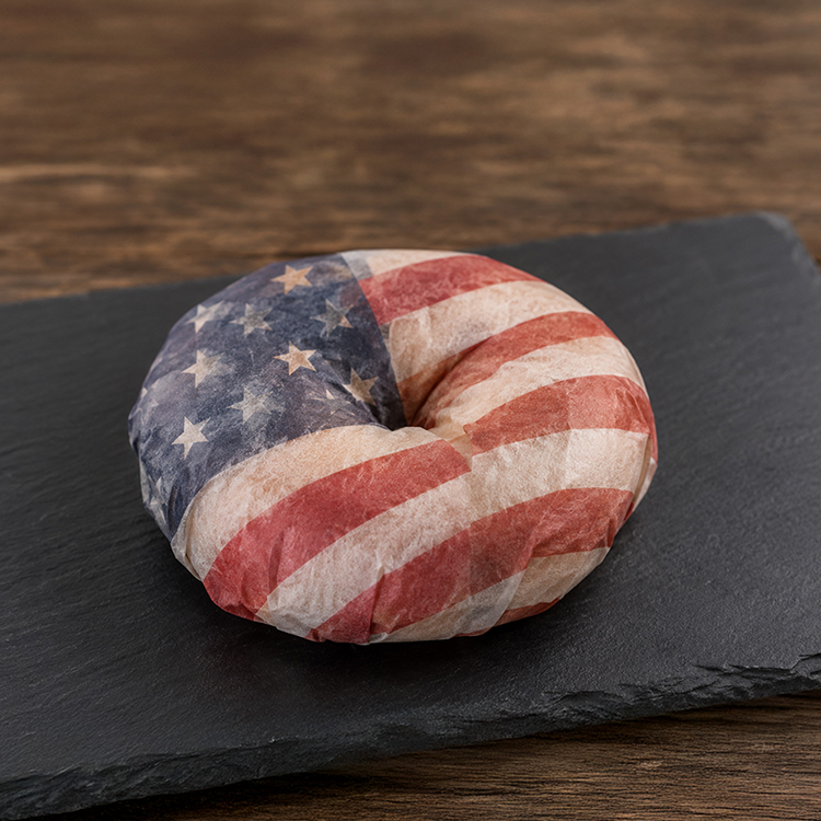 A bagel wrapped in paper printed with the American flag sits on a dark slate board—a true deli treat that invites you on a culinary journey around the world right at your table.