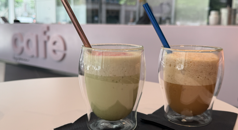 Zwei doppelwandige Gläser mit schaumigen Sommerdrinks - ein Eiskaffee und ein Matcha -, jeweils mit einem Strohhalm, stehen auf schwarzen Servietten auf einem weißen Tisch in einem modernen Café, dessen Schriftzug "Cafe" im Hintergrund verschwimmt.