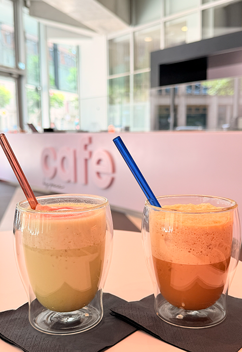 Zwei schaumige Sommerdrinks - ein leuchtender Matcha, der andere Eiskaffee - stehen in doppelwandigen Gläsern mit roten und blauen Strohhalmen auf schwarzen Servietten und erhellen ein modernes Café mit großen Fenstern und einer schlichten weißen Theke.