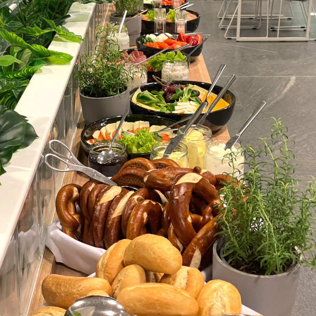 Ein Buffettisch mit frischen Brezeln, Brötchen, Salatschüsseln, Gemüse, verschiedenen Dips und Kräutertöpfen, ordentlich angerichtet in einem modernen Innenraum.