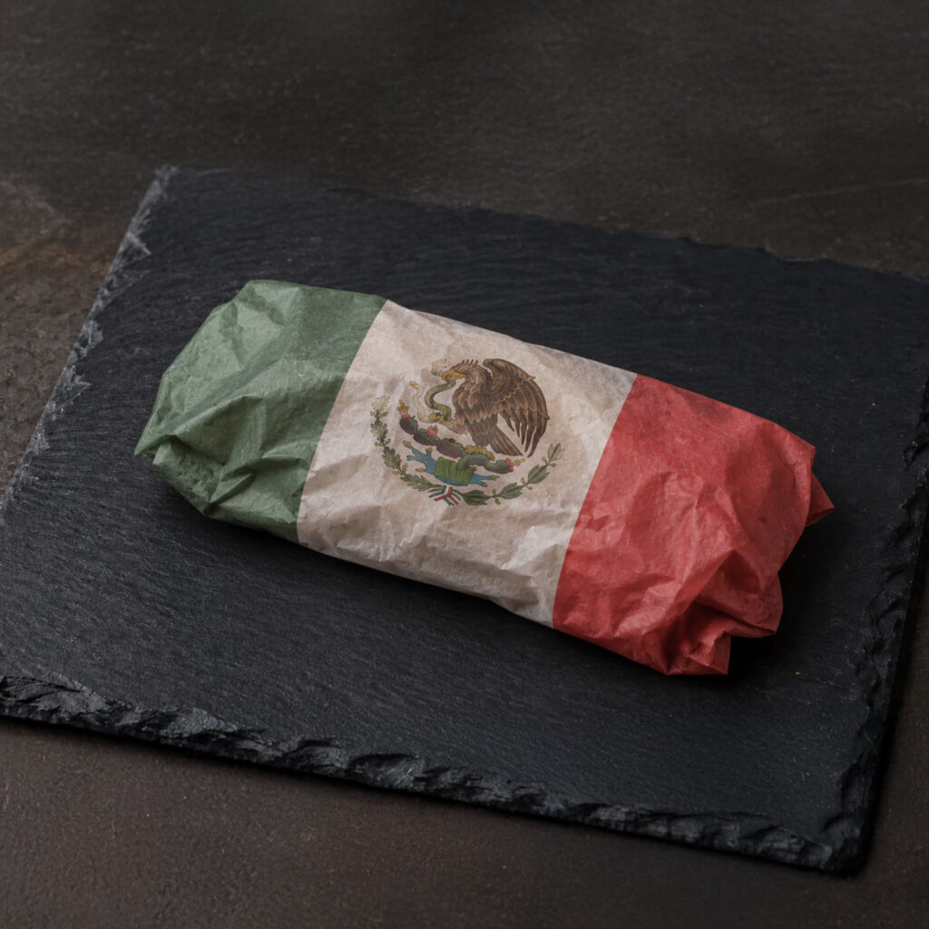 A wrapped burrito on a dark slate plate, with wrapping paper designed to resemble the Mexican flag—featuring green, white, and red stripes and the national coat of arms in the center.