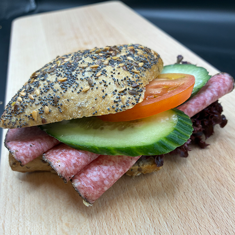 Auf einem Holzschneidebrett liegt ein Sandwich mit Mohnbrot, Salamischeiben, Gurke, Tomate und Salat.