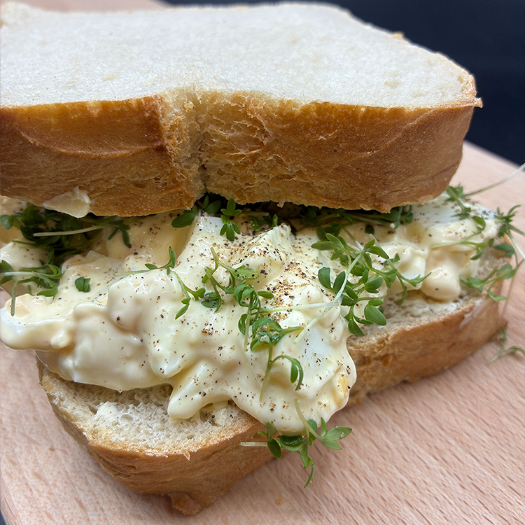 Eine Nahaufnahme eines Eiersalat-Sandwichs mit Mayonnaise und frischen Kräutern auf dicken Scheiben Weißbrot, das auf einer Holzunterlage liegt.