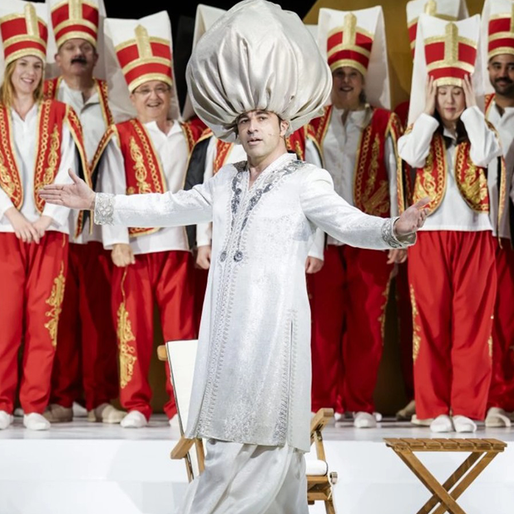 Ein Mann in einem aufwendig verzierten weißen Kostüm und einem hoch aufragenden silbernen Turban steht mit ausgestreckten Armen auf der Bühne der Staatsoper und tritt auf, während eine Gruppe in roten Hosen und hohen Hüten ihm zusieht – live im Livestream von BILD.de.