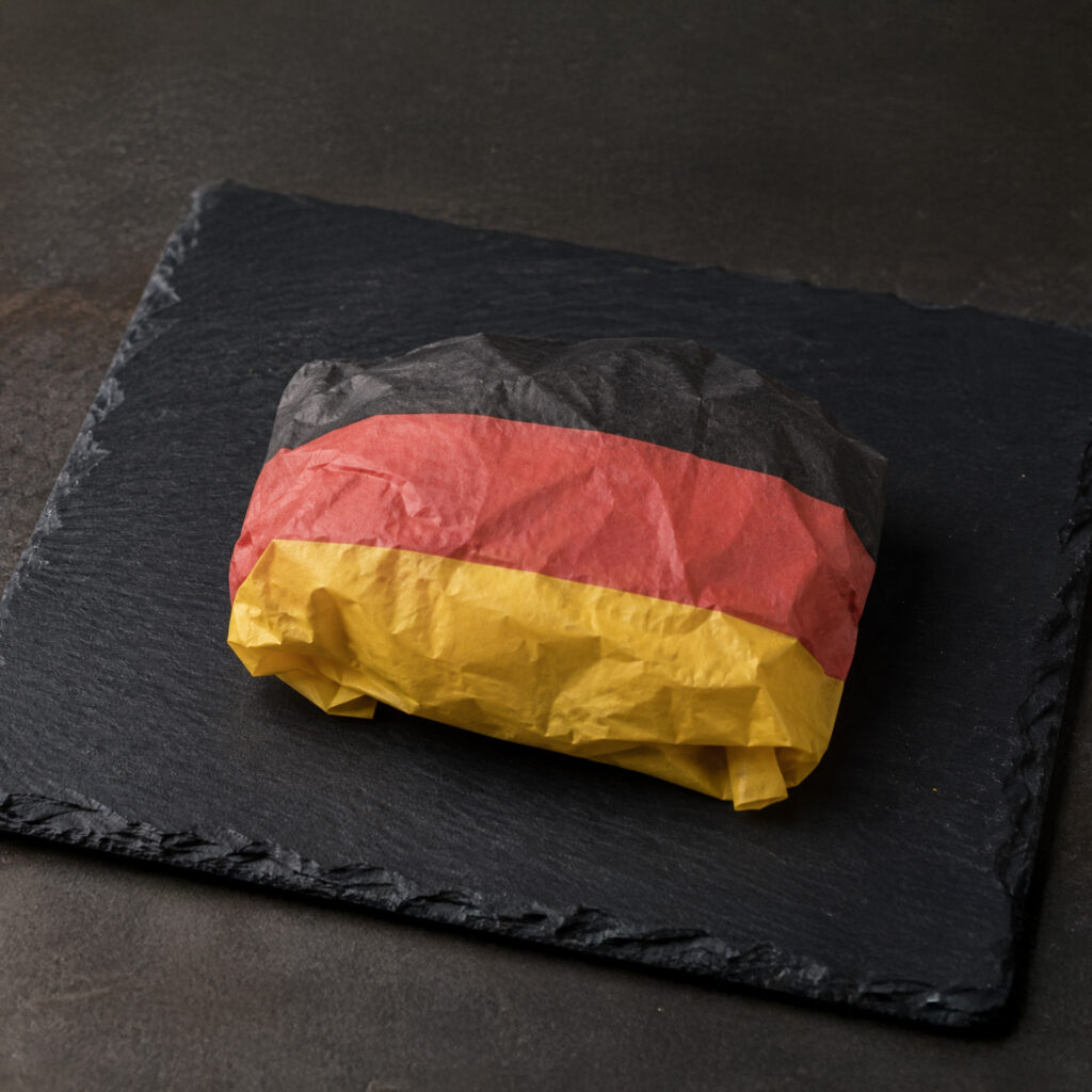 Ein Sandwich, eingewickelt in Papier in den Farben der deutschen Flagge – schwarz, rot und gelb –, liegt auf einem schwarzen Schiefer-Servierteller vor einem dunklen Hintergrund.