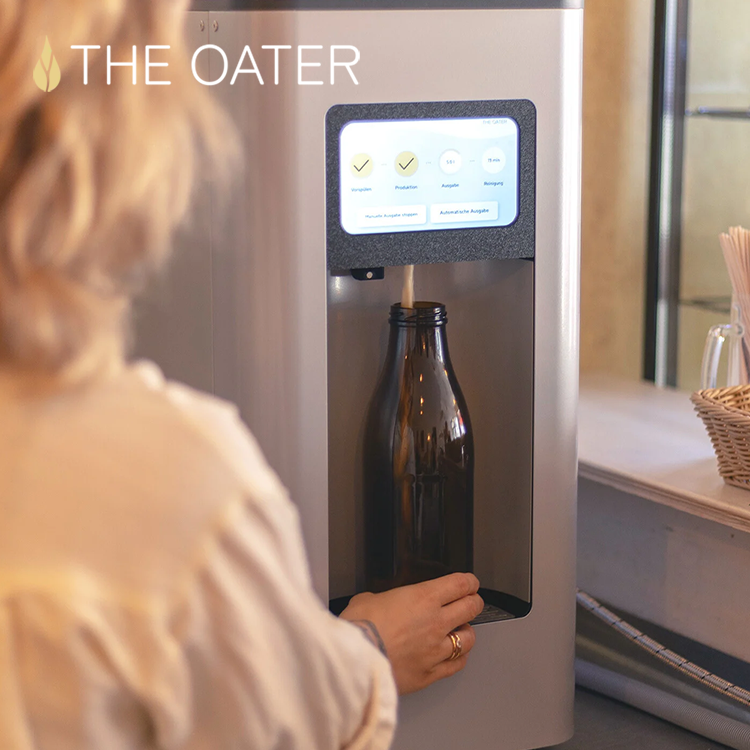 Eine Person füllt eine Glasflasche mit Hilfe einer Maschine namens „THE OATER“, deren digitales Display Optionen und den Betriebsstatus anzeigt – was an die moderne Technik erinnert, die in einem BILD.de-Livestream aus der Staatsoper zu sehen war. Die Maschine steht auf einer Arbeitsplatte neben einem Weidenkörbchen mit Servietten.