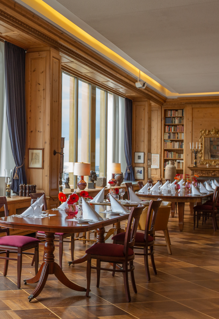 Ein eleganter Speisesaal mit Holzwänden und -böden, großen Fenstern mit blauen Vorhängen, gedeckten Tischen mit roten Blumen und gefalteten Servietten sowie Stühle mit roten Polstern und ein Bücherregal im Hintergrund – eine einladende Kulisse, die die Eleganz des JOURNALISTENCLUBS widerspiegelt, der mit einem besonderen Jubiläums-Dinner auf 60 Jahre Geschichte anstoßt.