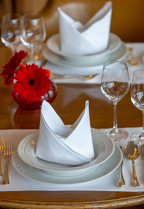Eine elegante Tischdekoration mit weißen Tellern, gefalteten weißen Servietten, Besteck, leeren Weingläsern und einem kleinen roten Blumenarrangement in einer runden Vase auf einem Holztisch – perfekt für ein Jubiläumsdinner zur Feier von 60 Jahren Geschichte im Journalistenclub.