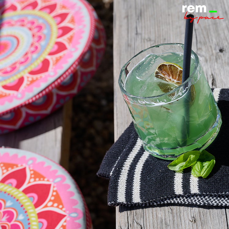 Ein grüner Cocktail mit Eis, einer Limettenscheibe und einem schwarzen Strohhalm steht auf einer gestreiften Serviette neben bunten Kissen mit Mandala-Mustern – perfekt, um sich nach dem Saison-Endspurt bei der pulsierenden Energie von DIE REM zu entspannen.