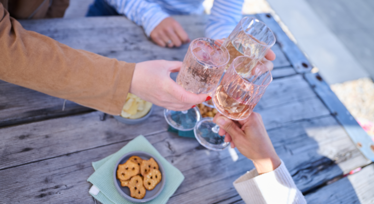 Drei Menschen stoßen bei Tageslicht an einem rustikalen Holztisch mit Snacks wie Brezeln und Chips mit ihren Gläsern Rosé an – und feiern den Saison-Endspurt mit viel Gelächter und in guter Gesellschaft.