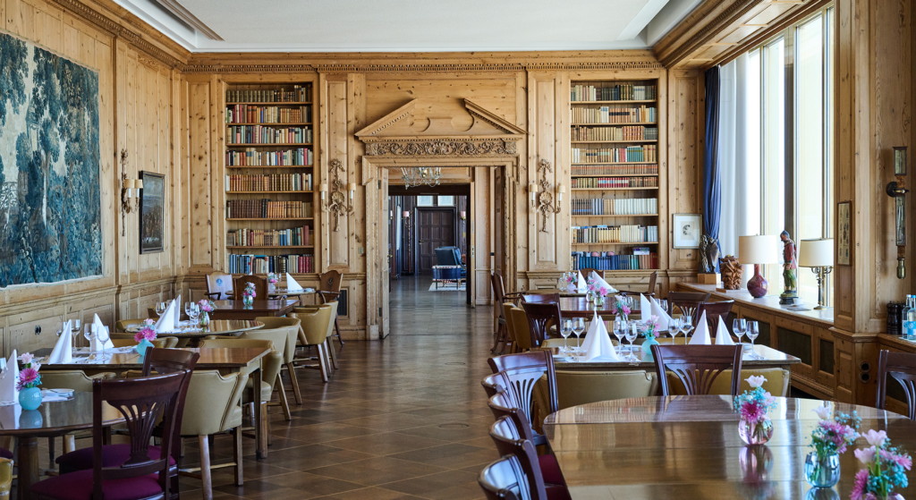 Ein mit Holz vertäfeltes Esszimmer mit gedeckten Tischen, Blumengestecken, Bücherregalen, großen Fenstern mit blauen Vorhängen und einer Tür, die in einen anderen Raum führt – ein elegantes Ambiente, perfekt für ein Jubiläumsdinner des JOURNALISTENCLUBs zur Feier von 60 Jahren Geschichte.