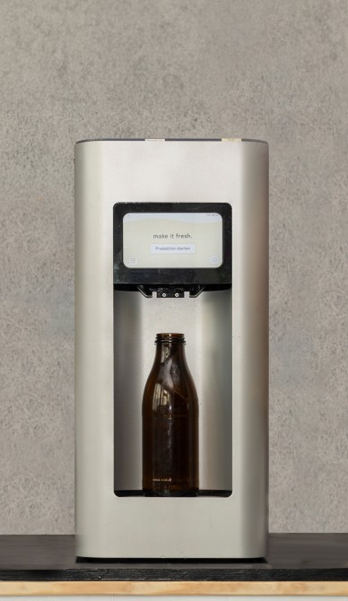 A stainless-steel beverage dispenser is filling a brown glass bottle with “Fresh Oats,” while text appears on a digital screen above it. A plain, light-colored wall is visible in the background.