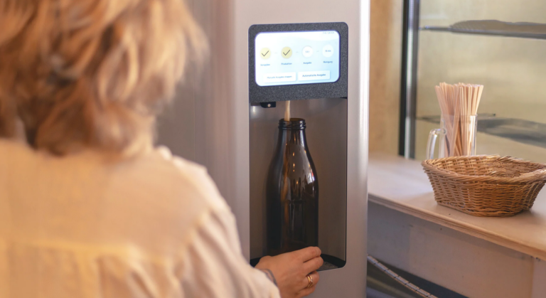 Jemand füllt an einem Wasserspender mit digitalem Bildschirm eine Glasflasche und hält dabei vielleicht kurz inne, um die neuesten Meldungen im BILD.de-Livestream zur „Oper über den Dächern Berlins“ zu verfolgen. Auf der Theke daneben steht ein Korb mit Rührstäbchen aus Holz.