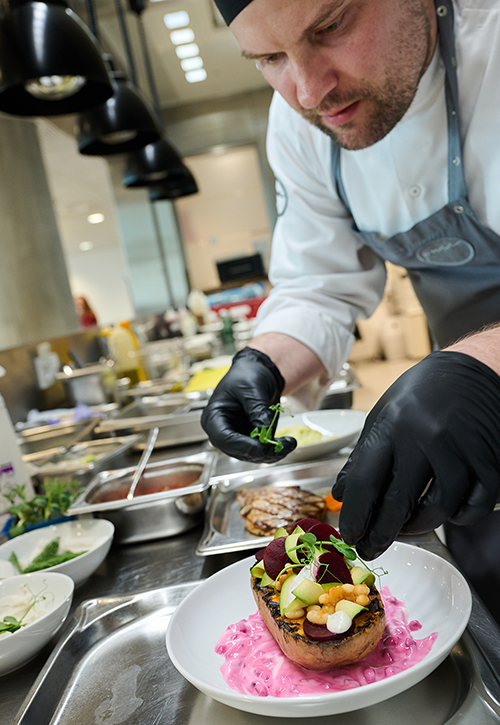 In a professional cafeteria kitchen, a chef wearing black gloves carefully plates a colorful dish on a white plate with leafy greens, surrounded by various ingredients and plates, offering a glimpse behind the scenes of the kitchen.
