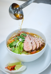 Using a ladle, broth is poured into a bowl of noodle soup garnished with slices of meat, vegetables, and herbs— a comforting favorite in any cafeteria. On the plate, lime, chili, and a crispy chip await as side dishes, adding extra flavor to every bite. Discover more insights into the kitchen as you savor every spoonful of this classic dish.