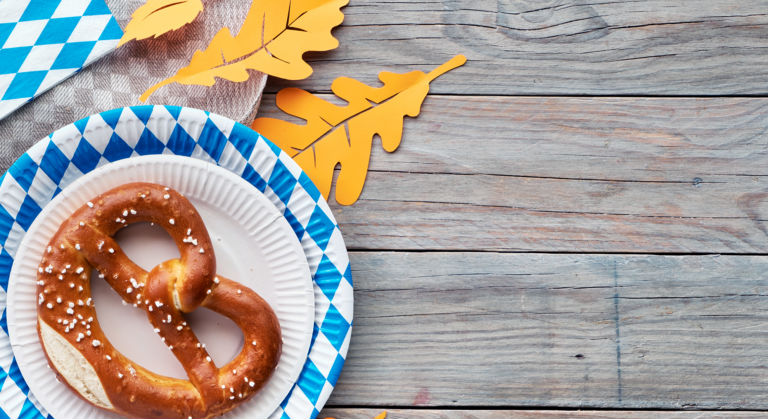 Eine gesalzene weiche Brezel auf einem weißen Teller, dazu eine blau karierte Serviette, gelbe Papierblätter und ein Holztisch im Hintergrund – das alles schafft eine einladende Oktoberfest- oder Wiesn-Atmosphäre, die sich perfekt eignet, um den Herbst zu feiern.