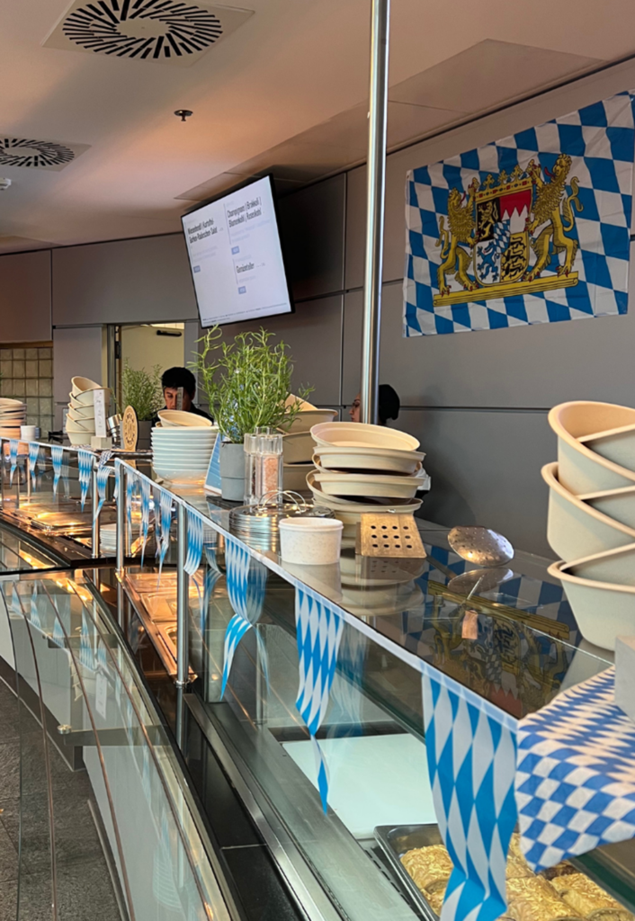 A cafeteria buffet featuring Bavarian blue-and-white flags, a Bavarian coat of arms, stacked plates, herbs, and serving trays conveys the festive spirit of Oktoberfest. In the background, a digital menu and two people can be seen, adding to the lively Wiesn atmosphere.