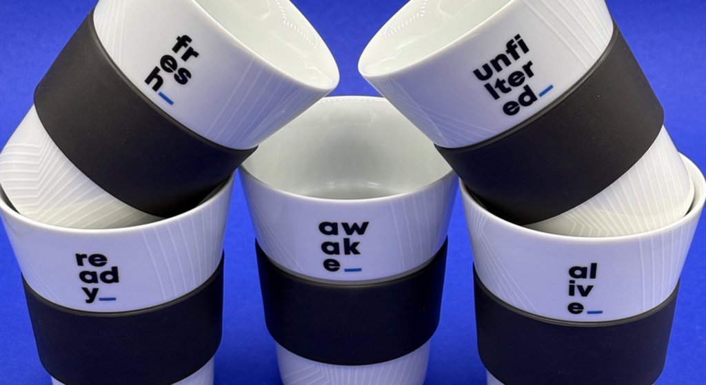 Fünf weiße Keramik-Kaffeetassen mit schwarzen Silikonhüllen für Kaffeeliebhaber, jede mit einem anderen Wort bedruckt: frisch, ungefiltert, bereit, wach und lebendig.